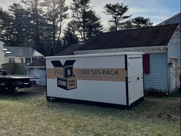 A container on a lawn beside a garage, a flatbed trailer parked at the road.