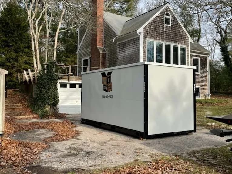 A container on a gravel drive beside a shingled house, bare trees overhead.