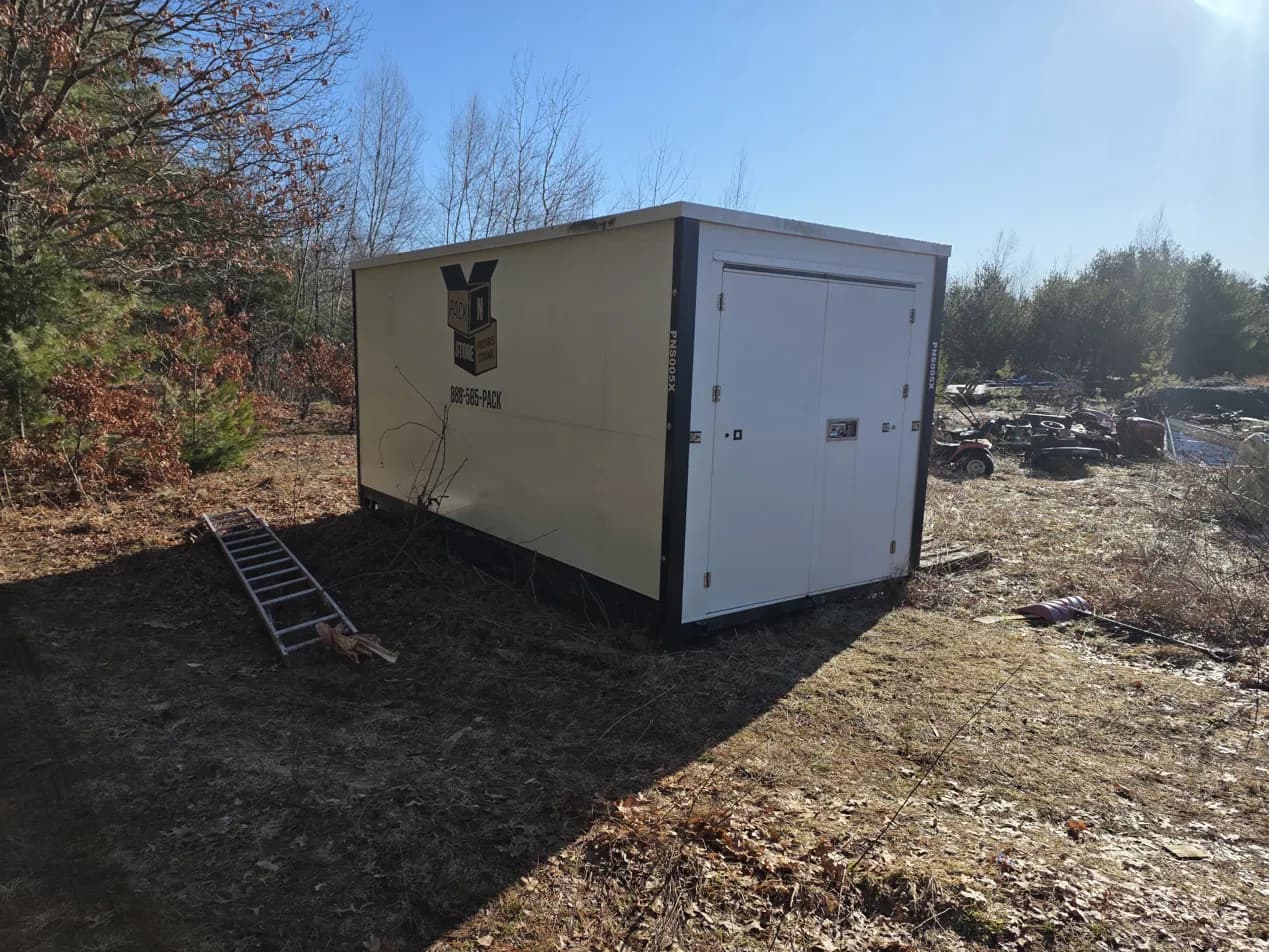 A container on scrub ground at the edge of a treeline, an aluminium ramp lying beside it, bare trees and blue sky behind.