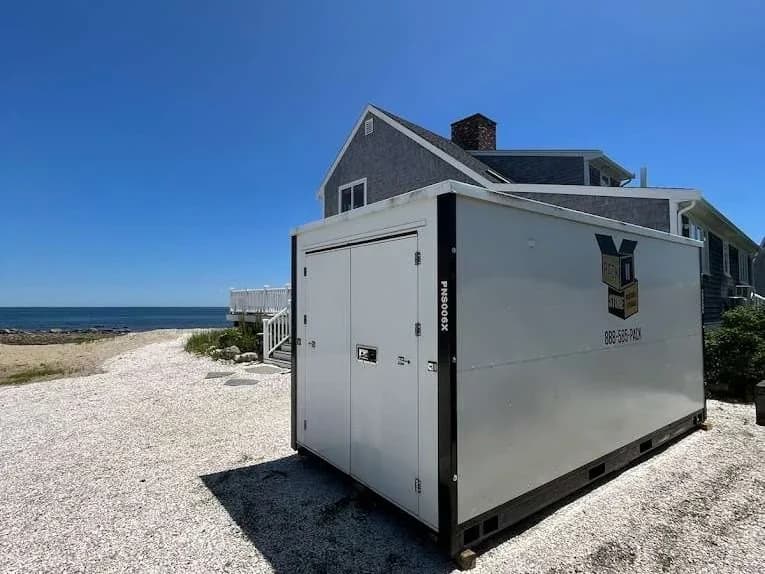 A container on crushed shell beside a shingled house, open water on the horizon.