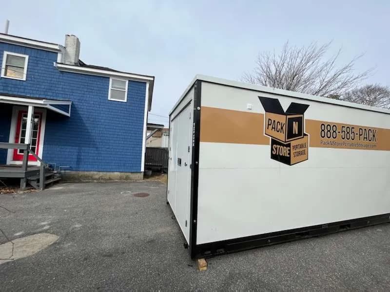 A container on a paved drive beside a blue clapboard house, plank under the corner.