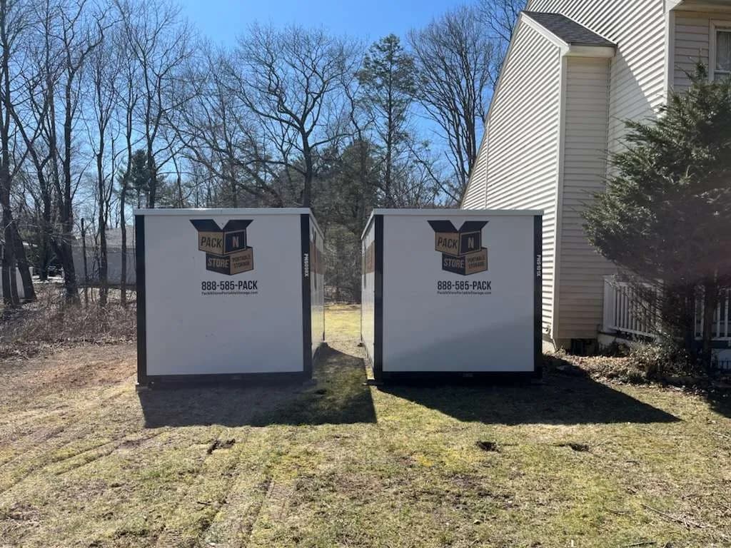 Two containers side by side on a lawn beside a townhouse, tyre ruts in the grass.