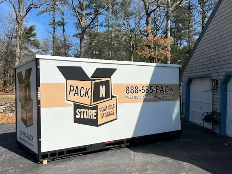 A container on a driveway beside a garage, planks under the corners.
