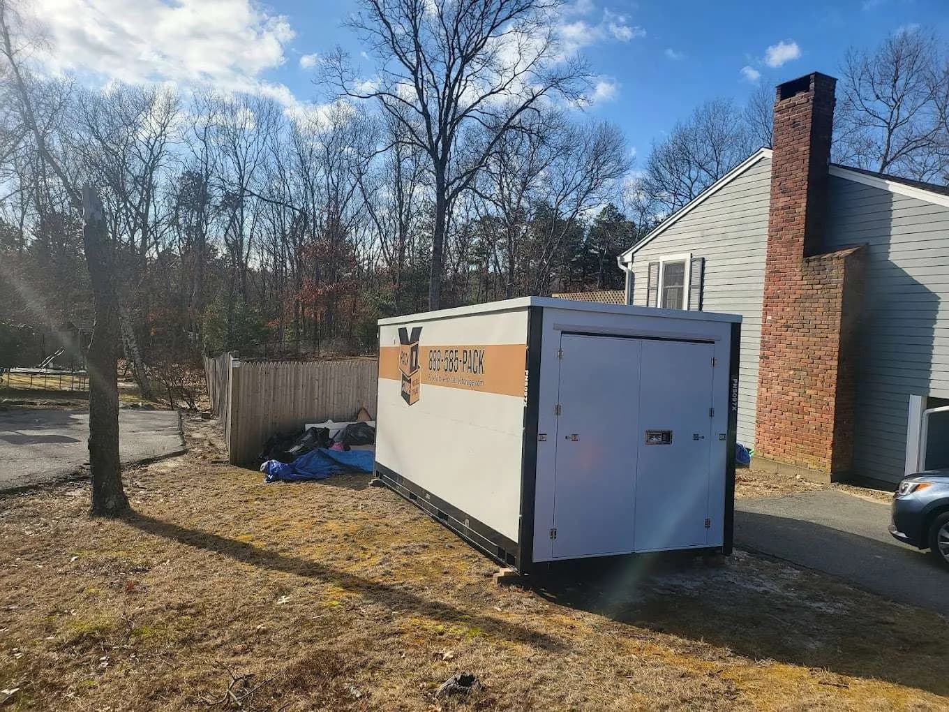 A container on packed grass between a chimney wall and a fence, doors facing the drive.