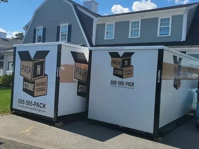 Two containers side by side on a driveway in front of a gambrel-roofed house.