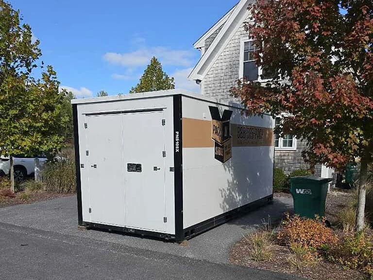 A container on pavement beside a shingled house, a tree in red fall leaves to its right.