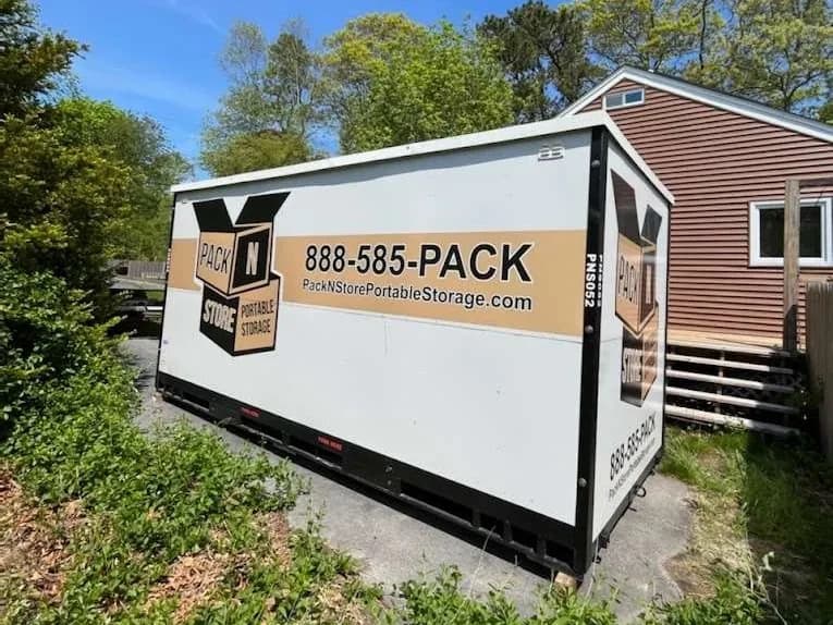 A container wedged between a house and a hedge on a narrow paved apron.