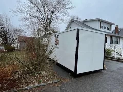 A container on a paved drive beside a house, heavy fog and bare trees behind.